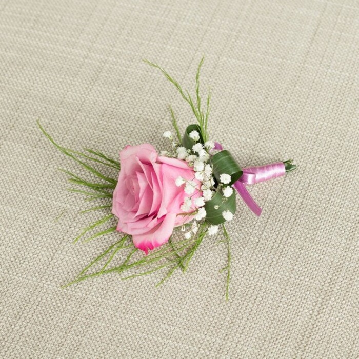 A finely crafted pink rose boutonniere rests on a light beige, linen-like fabric, whose subtle texture and gentle creases provide a warm, understated backdrop reminiscent of a carefully laid wedding table in a Mill Hill venue. Photographed from directly above, the design centres on a single, well-formed rose, its petals arranged in tight inner spirals that gradually relax into broader, open layers at the outer edge. The colour is a graceful gradient, beginning as a soft, blush pink at the core and deepening to a richer, more saturated rose tone at the tips, suggesting both freshness and tender romance. Clustered snugly around the base of the bloom are sprays of white baby's breath, each minute, rounded flower contributing to a light, cloud-like collar that softens the transition between rose and greenery. From beneath these delicate accents, fine strands of bright green, grass-like foliage fan outward, lending a sense of movement and natural ease, much like the grasses found along the paths of Mill Hill Park on a sunny afternoon. A broader, darker green leaf is neatly folded and wrapped at the base, encasing the stems in a tidy, architectural line that gives the piece a tailored, almost couture finish. The stems themselves are bound with a slim pink satin ribbon, tied into a compact bow; the ribbon's soft sheen catches the light, adding an elegant highlight without overpowering the flowers. Gentle, even lighting creates minimal shadow, allowing each petal, leaf, and sprig of baby's breath to be clearly defined. This image captures the quiet beauty of a classic rose boutonniere, the kind a groom or prom guest in Mill Hill might choose for a refined yet romantic touch, whether for an intimate gathering at home or a formal celebration nearby.