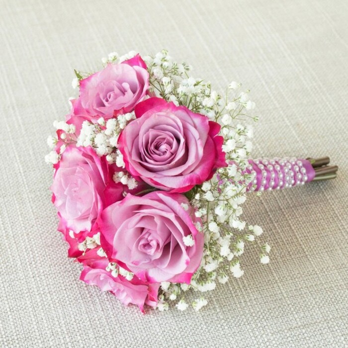 This arresting wedding bouquet, perfectly displayed atop a linen backdrop the shade of Mill Hill's gentle morning mist, is a testament to floral excellence and occasion-worthy artistry. Five large roses feature at its centre, their petals unfolding from deep, rich fuchsia-pink at the rim into soft, cherished lavender towards the inner core-a colour story as nuanced as Mill Hill's own community legacy. Between these lush blooms twist sprays of fine, pristine gypsophila, providing feathery contrast and an impression of buoyancy and grace. Each stem is expertly wrapped in regal purple satin, the handle encircled by several delicate strands of iridescent white pearls spiralling in harmony, reminiscent of celebratory toasts at local events on Bunns Lane. The natural ends of the stems peek out beneath, confirming the freshness of the flowers and the authenticity of the arrangement. Bathed in natural light, the bouquet is crafted in a round, full silhouette, its textures and tones revealed in all their subtlety. Ideal as a bridesmaid's bouquet or as a statement piece at a bridal shower along the leafy lanes of Mill Hill, this creation offers inspiration for elegant gatherings, evoking the romance and sophistication that define ceremonies in this North London suburb. The artistry echoes the expertise and pride of seasoned local florists, ensuring every petal plays its part in life's most memorable moments.