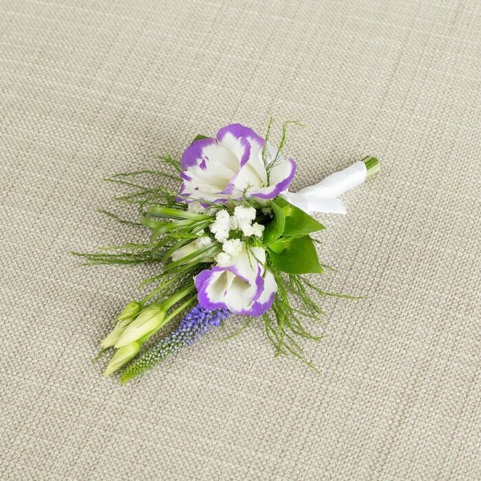 This image captures a refined boutonniere laid carefully on a pale beige, finely textured linen surface, the neutral background enhancing every nuance of colour and form in the flowers. Two lisianthus blooms dominate the composition, their petals a luminous white with dramatic, ruffled margins dipped in saturated purple, giving the impression of carefully inked edges on delicate parchment. Tucked between them are dainty clusters of white filler flowers, reminiscent of baby's breath, which break up the stronger tones and lend an airy, almost ethereal softness. A slender spire of muscari, or grape hyacinth, rises from the cluster, its tight, bead-like cups a deep violet-blue that brings a cool, textural contrast and evokes the feeling of an early spring walk through the gardens near Mill Hill Village. Around the flowers, fine, feathery greenery similar to asparagus fern spreads in light, arching fronds, providing movement and a slightly wild, garden-picked charm that balances the boutonniere's formal purpose. Two pale green buds rest near the edge of the design, still closed, embodying freshness and the sense that the floral story is not yet finished. The stems are trimmed, aligned, and wrapped in a smooth band of white satin ribbon, the fabric snug against the gathered stems and tied into a neat, understated bow that clearly marks the piece as ready for a groom's or groomsman's lapel. Crafted in a style often requested at Flowers Mill Hill for weddings around Mill Hill Broadway and nearby country houses, the overall effect is elegant, composed, and quietly romantic, ideal for anyone who prefers their flowers to convey grace through detail rather than grandeur.