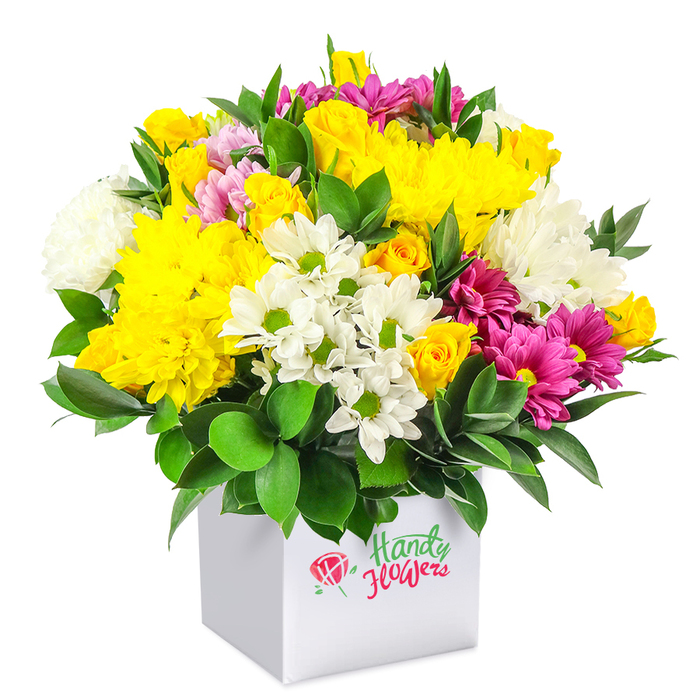 A vibrant mixed bouquet in a crisp white square box stands out dramatically against a clean white background, its dome-like shape suggesting abundance and care. The upper section of the arrangement is bathed in sunny yellow, dominated by large chrysanthemums whose densely packed petals form luxuriously full blooms. Nearby, small yellow spray roses gather in tight clusters, some buds still smooth and closed, others gently opening to reveal spirals of soft petals, evoking tenderness and hope. Among these golden tones, white chrysanthemums and daisy-like blooms with bright green centres weave in and out, their snowy petals reflecting the light and bringing a sense of calm and clarity. Cutting through this harmony are fuchsia and magenta chrysanthemums, sprinkled throughout the bouquet like scattered jewels; a few show lighter pink accents at the tips, creating a gentle gradient that draws the eye from one blossom to the next. A collar of deep green, glossy foliage surrounds the base, sending small sprays upward between the flowers, reminiscent of the well-tended gardens and leafy avenues around Mill Hill. The container, a smooth-sided cube-shaped white box, offers a modern alternative to a traditional vase and makes the bouquet easy to display on a windowsill overlooking Mill Hill Park or a desk near Mill Hill East station. The even lighting accentuates every fold of petal and shine of leaf, suggesting freshly arranged stems, cool to the touch and subtly fragrant. Overall, the composition conveys cheerful sophistication, suited to birthdays, anniversaries, or simply bringing a splash of fresh colour into a home or office across NW7, with the quiet assurance of quality associated with Flowers Mill Hill.