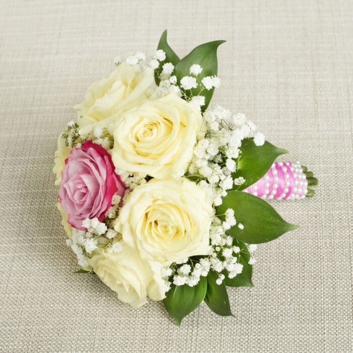 From a gentle overhead angle, this meticulously composed bouquet celebrates Mill Hill's refined aesthetic and the touch of tradition cherished in local gatherings. Plump, creamy roses-five or six in number-are arranged compactly, their petals mostly furled, signaling freshness and anticipation. They are anchored by a single, bold fuchsia-pink rose, which stands as a vibrant counterpoint among the soft pastels, drawing the eye like a wildflower in the meadows of North London. Lacy clouds of white baby's breath interlace throughout the roses, their texture as ethereal as morning fog drifting over the greenspaces of Mill Hill. Framing the bouquet, lush dark leaves provide a polished outline and a natural lift to the composition. The visible handle is tightly wrapped in luxurious fuchsia-pink satin ribbon and encircled by delicate strings of white pearls, their sheen offering glamour suited for elegant Mill Hill celebrations. Resting on a light, canvas-like fabric, the bouquet's colors and textures are illuminated by soft light, creating a sense of quiet joy and sophisticated romance for weddings, anniversaries, or community occasions.