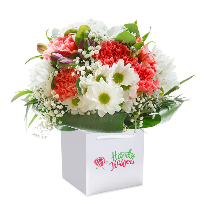 Brimming with colour and refined detail, this hand-tied bouquet is presented upright in a flawless, square white gift bag, the hallmark of an expertly curated selection from Flowers Mill Hill. An exuberant array of frilly carnations, ranging in hue from blush peach to lively coral and rich red, fills the arrangement's core-their ruffled petals catching the light and creating visual depth. Punctuating the bouquet, crisp white daisy-chrysanthemums with vibrant lime-green eyes lend a sense of joyful clarity, evoking memories of local gardens and the gentle openness of Mill Hill's green spaces. The soft movement of baby's breath, arranged in light clusters, brings a delicate texture and softness that contrasts gently with the fullness of the main blooms. At the left edge, touches of closed, purple-tipped buds add subtle dimension, suggesting new beginnings and unexpected delights. Encircling the flowers, broad, glossy green leaves and sprigs of fresh foliage offer structure and frame, holding the bouquet in a graceful, rounded silhouette. The white gift bag, set on a pristine white backdrop, accentuates the arrangement's vibrancy, while strong ribbon handles reinforce its suitability for easy gifting around the NW7 area-whether for a milestone celebration in Mill Hill Village or a compassionate gesture to a neighbour. The entire composition radiates freshness, artistry, and attentive local craftsmanship, making it an exemplary gift for any cherished occasion.