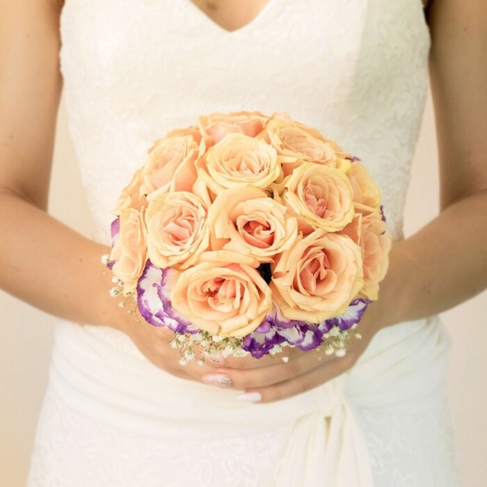 A refined, close-up photograph focuses on the midsection of a bride in a traditional ivory wedding dress, holding a round bouquet that becomes the clear focal point of the composition. The lace bodice of her gown is intricately detailed, with delicate floral patterns and a soft V-neckline, while the fabric at the waist is subtly ruched, giving way to gently cascading folds of skirt that fade into the background. Her hands, positioned at chest level, cradle the bouquet with a sense of care and expectation, nails neatly manicured in a pale nude shade with a faint glitter accent on one thumb, adding a whisper of sparkle that catches the light. The bouquet itself is an artful study in warm pastel colour, crafted as if by a seasoned florist accustomed to weddings across Mill Hill's chapels and garden venues. At the centre, densely clustered roses in shades of soft peach, apricot and blush pink create a plush, domed surface, each bloom perfectly open yet still slightly cupped, their velvety petals layered in tight spirals. This cloud of roses is framed by a contrasting collar of deep purple flowers edged with creamy white-likely lisianthus-whose ruffled petals add movement and a cool, sophisticated note around the base. Tiny, star-like sprays of white baby's breath are tucked in at intervals, softening edges and lending an airy, romantic halo to the design. The lighting is gentle and even, with no harsh shadows, suggesting an indoor setting just before the ceremony begins, perhaps in a quiet room overlooking the leafy streets of Mill Hill. The overall effect is one of elegance, tenderness and poised celebration, capturing the bouquet at the precise moment before it accompanies the bride down the aisle.