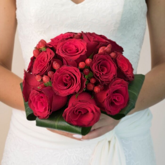 In this artful composition, a bride-moments from her Mill Hill ceremony-holds a lavish, jewel-toned bouquet as a centerpiece to the day. Her classic white wedding dress, adorned with delicate lace and a softly draping skirt, evokes the timeless grace often seen at local events along Daws Lane or in Mill Hill Village. The bouquet is a study in passion and luxury: a dense collection of velvety, rich crimson roses, each bloom pressed tightly to its neighbour, their intricate spiral centers and layered petals catching the soft light. Dotted throughout are clusters of vibrant red hypericum berries, providing a sparkling, textural counterpoint to the unified reds of the roses. The bouquet's base is structured by a collar of neatly folded, deep green leaves-glossy, bold, and reminiscent of Mill Hill's leafy avenues. The overall effect, illuminated by even lighting against a gently blurred and neutral background, is ethereal and enchanting, highlighting the wordless emotion and careful craftsmanship. This arrangement, available for delivery to Mill Hill and surrounding neighbourhoods, epitomises the elegance and celebration of North London weddings, providing both fragrance and colour to a day filled with meaning.