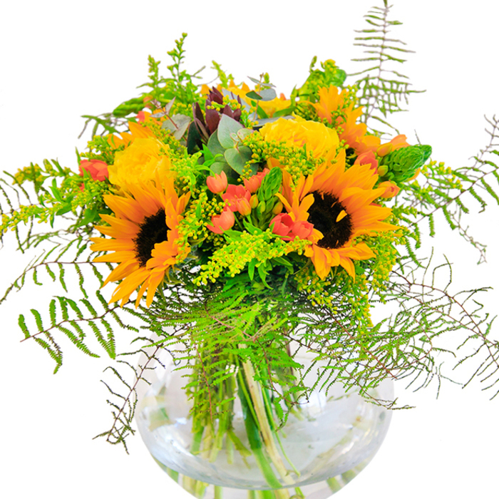 The overwhelming sense from this bouquet is joy and vitality, as if Mill Hill's open spaces have been gathered into a glass vase. Towering sunflowers with dark, earthy centers and golden petals form the heart of the design, their cheerful presence impossible to miss. They are joined by neatly furled yellow garden roses, whose velvet roses spiral outwards with understated elegance, softly perfumed and delicately colored. Ruby-red hypericum berries nestle throughout, drawing the eye and creating a playful dialogue with the surrounding greenery. The arrangement is alive with motion-cascading fronds of fern, wispy fillers of solidago with light yellow florets, and the cool shimmer of eucalyptus leaves that hint at distant fields. The clear, globe-shaped vase, reminiscent of those seen in stylish Mill Hill cafes, holds every stem in pure water, magnifying shades and forms while keeping the composition fresh and bright. Peer close and you'll find a textural interplay: the sunflowers' plush suede, crisp green leaves, and the smooth, glossy finish of the berries in perfect balance. Designed as a centerpiece for birthdays, anniversaries, and all those moments where happiness takes center stage, this bouquet honors the natural beauty found across Mill Hill's green heart and is expertly crafted by Flowers Mill Hill for local celebrations.