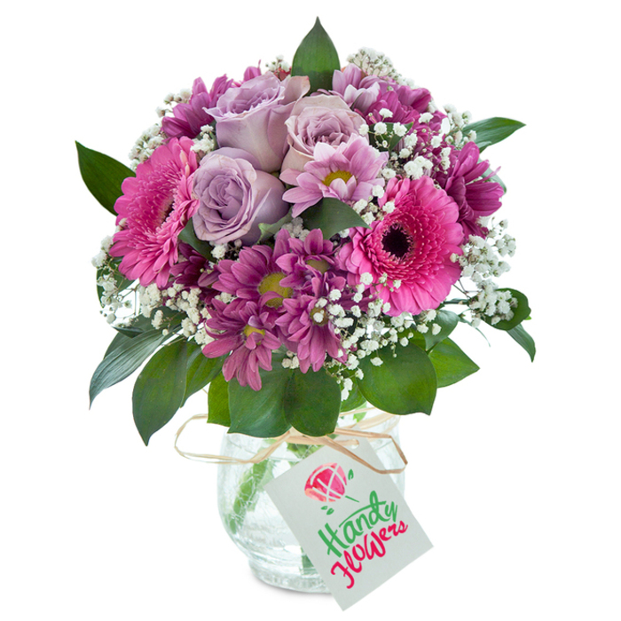 Perfectly arranged in a clear glass vase with a charming crackled finish, this mixed bouquet from a Mill Hill florist is a study in colour, harmony, and freshness. At its heart, several lavender roses bloom softly, the gentle unfurling of their petals reminiscent of delicate mornings in Mill Hill's Finchley Road gardens. Encircling the roses are vivacious Gerbera daisies in radiant fuchsia pink, their unmistakably bold centers drawing the eye and lifting the mood, like sunshine breaking through clouds over Mill Hill Village Green. Deep magenta chrysanthemums, their petals plush and vivid, cluster throughout to intensify the arrangement's richness, while subtle hints of yellow-green in their hearts catch the light. Wisps of white baby's breath float among the more dramatic flowers, infusing a sense of airiness and bringing soft illumination that refreshes the overall effect. Robust green leaves emerge beneath, providing grounding and a visual echo of Mill Hill's abundant leafy avenues. The bouquet sits against a crisp white background, making its vivid palette especially striking. Around the vase's neck is tied a rustic raffia bow, supporting a white tag bearing the phrase 'Happy Birthday' in bright red and green lettering, topped with a cheerful pink flower emblem-a finishing touch that feels both festive and personal. This thoughtfully gathered bouquet stands ready as a heartfelt birthday gift or a celebratory gesture, perfectly reflecting the style and care for which Mill Hill's florists are known, and is readily available for fast, local delivery throughout the community.