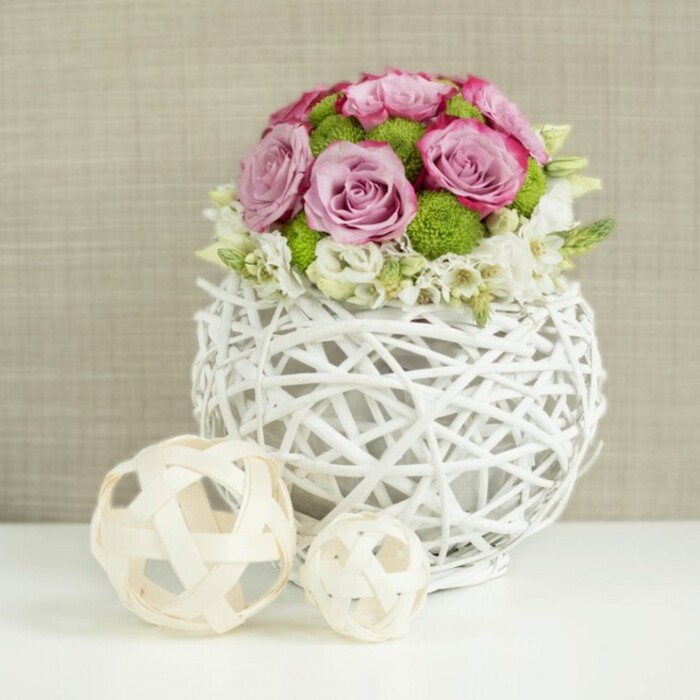 Captured in soft, even lighting, this floral arrangement feels like a quiet celebration ready to be placed on a wedding table in a Mill Hill garden marquee. The centerpiece is built as a rounded dome of blooms, overflowing from a distinctive spherical vase constructed from white, rattan-like twigs. These slender strands weave around one another in an open pattern, creating a basket-like globe with small gaps that lend an airy, contemporary feel. Within this cradle, roses in shades of blush pink and gentle lavender are clustered closely, many fully open, revealing layers of petals that spiral outward with a velvety sheen. Some petals are tinged at the edges with a slightly deeper pink, giving the flowers subtle depth and a sense of movement. Between the roses, bright green button chrysanthemums punctuate the design, their small, spherical heads packed with finely textured petals that bring a lively freshness reminiscent of Mill Hill's green spaces. At the base of the bouquet, delicate white filler flowers are interspersed among the larger blooms, softening the border where flowers meet vase and creating a gentle, almost cascading effect. On the crisp white surface to the left of the arrangement, two light wicker decorative balls lie side by side, one larger than the other. Their open, crisscross structure mirrors the vase and adds to the sense of carefully coordinated design. Behind everything, a softly blurred beige background, textured like linen or burlap, provides warmth and depth without distracting from the details. The overall impression is one of thoughtful craftsmanship and romantic elegance, perfectly suited to tablescapes across Mill Hill, from family homes to intimate local venues.