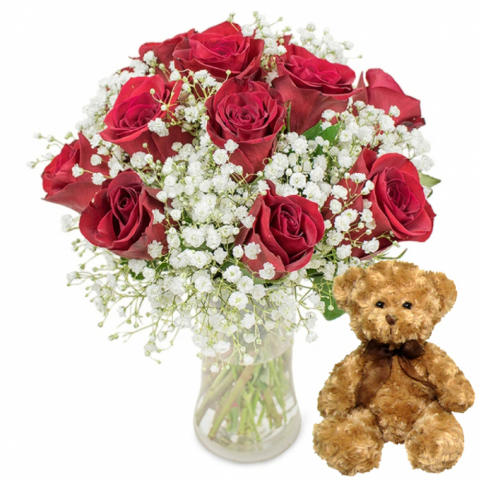 In this carefully arranged studio shot, a romantic duo of flowers and plush sits center stage, reminiscent of a thoughtful surprise being prepared in a Mill Hill florist's workshop. On the left stands a bouquet of around twelve deep red roses, proudly displayed in a clear glass vase filled with fresh water. Each rose is full and velvety, the petals curling into soft spirals that layer over one another like satin fabrics, creating a rich, textural depth. Some blossoms are more open, revealing their heart, while others remain partially cupped, lending a sense of movement and life to the arrangement. Nestled among the roses are clouds of white baby's breath, their tiny blossoms scattered along fine green stems, forming a light, frothy frame around the bolder crimson blooms. This interplay of intense red, bright white, and dark green suggests timeless romance, perfectly suited to the quieter, leafy character of Mill Hill, from the calm paths of Mill Hill Park to the nearby tree-lined avenues. The vase itself is simple and transparent, allowing the viewer to see the straight green stems inside, giving a sense of honesty and freshness. To the right of the flowers, a plush teddy bear sits upright on the same white surface. Its fur is light brown and softly textured, with rounded ears, a pair of glossy dark button eyes, a small stitched nose, and a gentle embroidered smile that conveys warmth and reassurance. A dark brown ribbon is tied neatly around its neck in a tidy bow, mirroring the sophistication of the roses. The clean, white background removes all distraction, focusing attention solely on this tender combination-a perfect visual symbol of love, apology, celebration, or appreciation for someone in Mill Hill, whether delivered to a home, an office near the station, or a cosy café rendezvous spot.