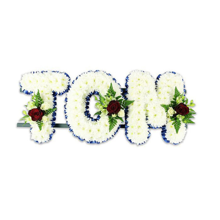 A carefully fashioned memorial spells out 'TOM' in substantial, three-dimensional letters made from countless small white chrysanthemums, their clustered petals forming a soft, tactile surface that reads as both pure and consoling. Each letter's perimeter is traced with a vivid royal blue ribbon, the colour acting as a crisp frame that accentuates the clean lines and adds a touch of quiet stateliness. Within the central field of each letter, a composed spray features a solitary deep red rose as its focal point - a classic symbol of enduring love - surrounded by pale ivory spray roses and lush green fern fronds which bring texture, depth and a gentle fragrance to the piece. A discreet dark-green or black metal framework is visible beneath the arrangement, providing stability so the letters can be presented upright at a funeral service or memorial display. Against the uncluttered white backdrop, the contrast between the snowy chrysanthemums, the royal-blue edging and the burgundy roses reads with clear, respectful intent. As a florist at Flowers Mill Hill who often arranges tributes for services at St Paul's or nearby chapels along The Ridgeway and Bittacy Rise, I design this type of tribute to balance simplicity with heartfelt detail-each bloom positioned with intention, each ribbon edge smoothed for a tidy finish. Prepared with fresh, seasonally-sourced flowers and offered for local delivery across Mill Hill, it's a bespoke sympathy arrangement that aims to honour a life with dignity and quiet beauty.