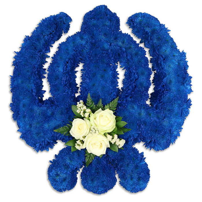 A beautifully crafted Sikh Khanda floral tribute dominates a stark white background, its bold royal blue structure instantly recognisable and rich with meaning. Every element of the symbol is carefully defined: the upright central khanda sword, the unbroken chakkar encircling it, and the two outward-sweeping kirpans that lend balance and protection. These forms are constructed from countless blue chrysanthemum florets, clustered so densely that they create a plush, cushion-like surface. From a distance the blue appears uniform, but on closer description, there are subtle shifts in tone within the petals, adding depth and a gentle sense of movement, as though a soft breeze were passing across the flowers. At the base of the central sword, a small, contrasting arrangement of white and green blooms becomes the emotional heart of the piece. Here, three pure white roses rest together, their petals smooth and delicately spiralled, holding the suggestion of fragrance and tenderness. Surrounding them are sprays of fine white filler flowers and a halo of dark green foliage that both frames and supports the roses, grounding the design in natural beauty. The lighting is calm and diffused, casting slight shadows beneath the petals and along the outer edges of the Khanda, giving it a three-dimensional presence that feels almost sculptural. Created with the expertise of a seasoned local florist, this tribute would sit beautifully at a Sikh funeral or remembrance service in Mill Hill, perhaps at a local place of worship or delivered quietly to a family home off Lawrence Street or near Mill Hill Broadway, as a respectful symbol of remembrance, devotion, and unbroken spiritual connection.