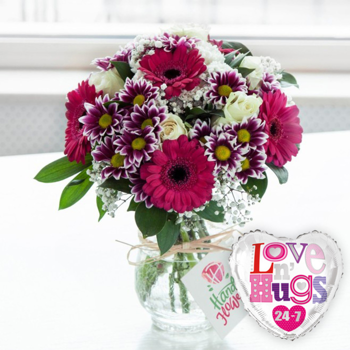 A celebratory floral gift is arranged on a pale surface, the kind of scene you might find in a cosy Mill Hill sitting room after a surprise delivery. The central focus is a clear, rounded glass vase brimming with a carefully balanced bouquet. Bold gerbera daisies in deep fuchsia and magenta dominate the front, their broad petals radiating around prominent, dark centres that lend a graphic, almost modern feel. Clustered around them, exquisite chrysanthemums catch the light with petals that shift from rich purple to clean white at the edges, each bloom brightened by a neat yellow centre, creating a lively contrast. Creamy white roses thread through the arrangement, their softly spiralling petals and gentle ivory tones adding an air of classic elegance. Delicate sprays of baby's breath, or gypsophila, float among the larger blossoms, sprinkling the bouquet with tiny white stars and softening the transitions between colours. Green foliage frames everything, with fresh, glossy leaves emerging from the base and sides, giving the design fullness and a natural, garden-like movement. The vase reveals the meticulously trimmed stems, all resting in clear water, their slightly angled lines meeting at the base in a sign of professional floristry. At the neck of the vase, a loop of rustic natural twine is tied neatly, holding a small white tag decorated with a simple pink rose and easy-going green script, as though hand-lettered just before delivery in Mill Hill. To the right of the vase, a shiny silver-edged heart-shaped foil balloon leans companionably toward the flowers, its white centre printed with the phrase Love n' Hugs 24-7 in a collage of pink, purple and blue patterned letters. The softly blurred, light-filled background suggests daylight shining through a nearby window, giving the entire composition a warm, affectionate glow suitable for birthdays, anniversaries or everyday gestures of love around Mill Hill Broadway.