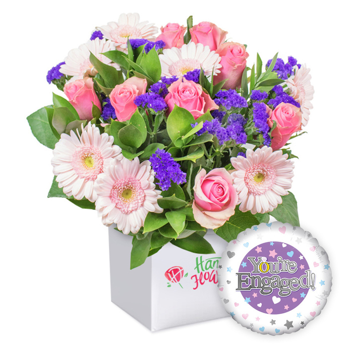 Elegantly arranged in a crisp white square box, this engagement bouquet radiates tender romance with contemporary flair, a signature offering for Mill Hill residents. The soft pink roses stand out, their petals unfurling in creamy spirals from tight buds to open blossoms, perfume light and sweet in the air. They mingle with pastel pink gerbera daisies, their white-edged petals haloed by pale yellow centers, creating a buoyant, sunlit mood. Interspersed among them are clusters of deep purple statice, lending depth and a whisper of mystery to the palette. A lush veil of green foliage weaves through the stems, providing a garden-fresh backdrop that evokes a quiet courtyard in Mill Hill East.

The blooms are neatly nestled in a clean, square white box, its front bearing a delicate red rose mark and green script that hints at local floristry craft without naming brands. In the lower right, a glossy foil balloon completes the gift: a round shape with a purple center bearing You're Engaged! in silvery type, and the golden 'O' styled as a ring. The outer white rim is dotted with pastel hearts and tiny silver stars, echoing the romance of the moment. This arrangement is a timeless congratulations for an engaged couple, available for dependable delivery around Mill Hill, Mill Hill East, and nearby streets, and well suited to same-day gifts that brighten a doorstep or office.