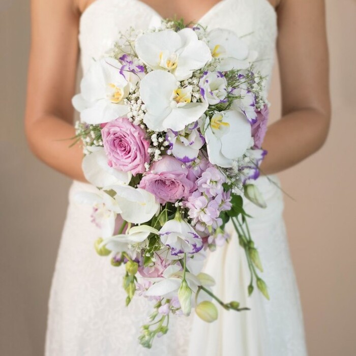 In a close-up tableau reminiscent of Mill Hill's own tranquil gardens, this image portrays a bride mid-frame, her torso gently curved around an extraordinary cascading bouquet-the true star of this elegant bridal composition. The soft, sunlit beige background imparts a luminous quality, helping each detail stand out: the wedding dress's lace bodice, with intricate patterns, appears just beneath the bouquet, its white expanse harmonizing with the array of flowers above. Dominating the bouquet are immaculate white phalaenopsis orchids, their sweeping petals and golden centers glowing amid pastel pink garden roses, each bloom meticulously chosen for fullness and texture. Lavender and pale purple lisianthus, with their unique fringed petals, are woven throughout, adding depth and a subtle lavender haze. Intersections of baby's breath create a diaphanous layer that lightens the structure, complemented by discreet strands of green foliage. The trailing, teardrop shape tapers gracefully, enlivened by unopened buds and slender vines, an echo of the rolling green expanses near Mill Hill Park. The overall design feels both contemporary and classic, ideally suited to brides seeking a blend of luxury and tradition within the NW7 postcode. The scene's artistry and gentle atmosphere exemplify the signature wedding flowers arranged locally by Flowers Mill Hill, capturing the emotion and promise of the day.