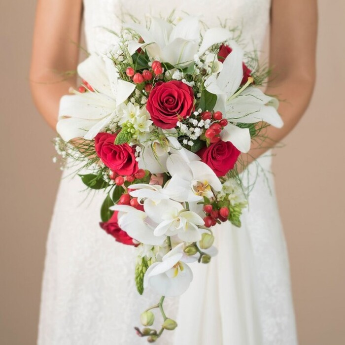 Lit by gentle daylight reminiscent of quiet Mill Hill mornings, this captivating bridal bouquet stands as a testament to refined floral artistry. The bride, presented from the torso upwards in a timeless white lace dress, offers the bouquet in both hands-a moment that radiates elegance just steps from the historic Ridgeway. Trumpet-like white lilies fill the majority of the arrangement with their opulent blossoms, their delicate stamens adding a sculptural, golden accent. Set against them, bold, rich red roses swirl with velvety layers, serving as passionate centrepieces, while the cascading effect is beautifully realised by long, arching stems of white Phalaenopsis orchids trailing to the bouquet's tip. Threaded between are lively bursts of hypericum berries, their deep crimson hues mirroring the vibrant offerings of Mill Hill farmers' stalls, and clusters of airy gypsophila provide a fragrant, cloud-like softness. Wispy green asparagus fern and other lush greenery establish the bouquet's natural shape, interspersed with an occasional spiky accent, adding dynamism and texture much like the undulating landscape of NW7. The warmly blurred background in subtle beige tones ensures every feature is on display, from the bride's gentle embrace to the precise placement of each bloom. With a harmonious blend of tradition and modern style, this exceptional bouquet from Flowers Mill Hill is designed for ceremonies reflecting love and North London charm.