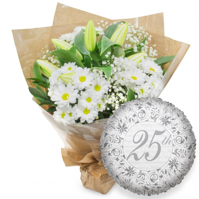The photograph captures a classic silver anniversary pairing: a fresh white floral bouquet and a beautifully detailed foil balloon, set against a spotless white background that accentuates their textures and tones. The bouquet is dominated by white chrysanthemum flowers, their many slender petals arranged like little suns around warm yellow centres. These chrysanthemums are closely packed, creating a plush, generous bed of white. Among them, elegant white lily buds rise up, long and tapering, with just a hint of green along their edges. Some buds are tightly closed, while others appear on the verge of opening, lending the arrangement a sense of life in motion. Delicate clusters of baby's breath weave between the main flowers, the tiny white blossoms forming a soft mist that lightens the overall feel. The entire arrangement is supported by rich green foliage, with leaves that curve outward from the base, giving depth and framing the blooms much as the leafy streets of Mill Hill frame its family homes. The bouquet is wrapped in a layer of clear cellophane that adds a slight shine, then covered in brown kraft paper folded carefully around the stems, evoking the look of market-fresh flowers carried from Mill Hill Broadway or along Lawrence Street. The base of the bouquet is neatly bound, ready to be presented by hand or delivered locally. To the lower right side, a round silver foil balloon sits partially overlapping the flowers. Its metallic surface shows gentle ripples and is adorned with intricate scrollwork and floral motifs printed in a darker silver. At its centre, the inscription 25th appears in graceful script, surrounded by a background pattern of softly repeated Happy Anniversary phrases in pale grey. Together, bouquet and balloon create an image of tasteful celebration, perfect for commemorating a Silver Anniversary in the Mill Hill area.