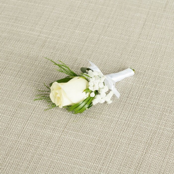 An elegant wedding boutonniere featuring a single white rose is laid diagonally on a pale beige, linen-textured surface, evoking the quiet preparation moments before a ceremony in Mill Hill's leafy streets. The rose itself is a tightly furled bud of creamy white, with petals so smooth and velvety they appear almost porcelain-like, cupping gently toward the centre as though just beginning to unfurl. At the base of the bloom, small hints of soft green sepals peek out, adding a natural transition between flower and stem. Surrounding the rose are light, airy clusters of baby's breath, each tiny white blossom forming part of a cloud-like surround that gives the piece a dreamy, ethereal softness. Interspersed among these are slender, vibrant green leaves and a few fine, fern-like sprigs, their lines clean and slightly arching, framing the focal bloom with a fresh, garden-inspired look typical of floristry in this North West London neighbourhood. The stems are carefully aligned and wrapped in a band of pristine white satin ribbon, smooth and slightly glossy under the light. Just beneath the floral heads, the ribbon is tied into a neat, graceful bow, the loops and trailing ends lying flat and controlled, suggesting the work of a meticulous florist preparing it for a groom's lapel. The textured background, with its subtle crosshatch weave, provides a neutral, organic setting that highlights every detail of the flowers and greenery without distraction. Soft, even lighting washes across the scene, enhancing the gentle shadows between petals and the depth of the foliage, creating a feeling of calm, polished sophistication. This image conveys the tenderness of wedding rituals in Mill Hill-from quiet homes near the Ridgeway to elegant venues nearby-capturing a single, refined symbol of love, tradition, and new beginnings.