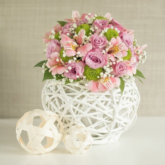 Bathed in soft daylight, this floral centerpiece becomes the focal point of an inviting Mill Hill gathering-its colors and textures echoing the refined traditions and modern touches of northwest London homes. The arrangement features a multitude of pastel pink and lavender roses in full bloom, petals unfurling in plush, layered rosettes suggestive of a summer walk through Mill Hill Park at golden hour. Between these, gentle pink alstroemeria stand out with slender petals and subtle inner markings, while clusters of lime green button chrysanthemums add animated pops of color and compact, tactile interest. Wisps of delicate baby's breath thread throughout, imparting an airy, ethereal feel complemented by deep green foliage poking through to ground the bouquet in fresh natural vibrance. The flowers spill from an eye-catching white vase, its open-weave texture reminiscent of expertly woven baskets spotted at local community shops, its rounded form lending stability. In the foreground, two decorative spheres in natural, woven textures echo the intricate vase and reinforce the arrangement's handcrafted theme, their placement along the crisp white table reminiscent of artful Mill Hill interiors. With a backdrop of warm, neutral beige, the centerpiece projects sophistication, freshness, and understated joy, making it an ideal choice for weddings, anniversaries, or elegant entertaining within the Mill Hill community.