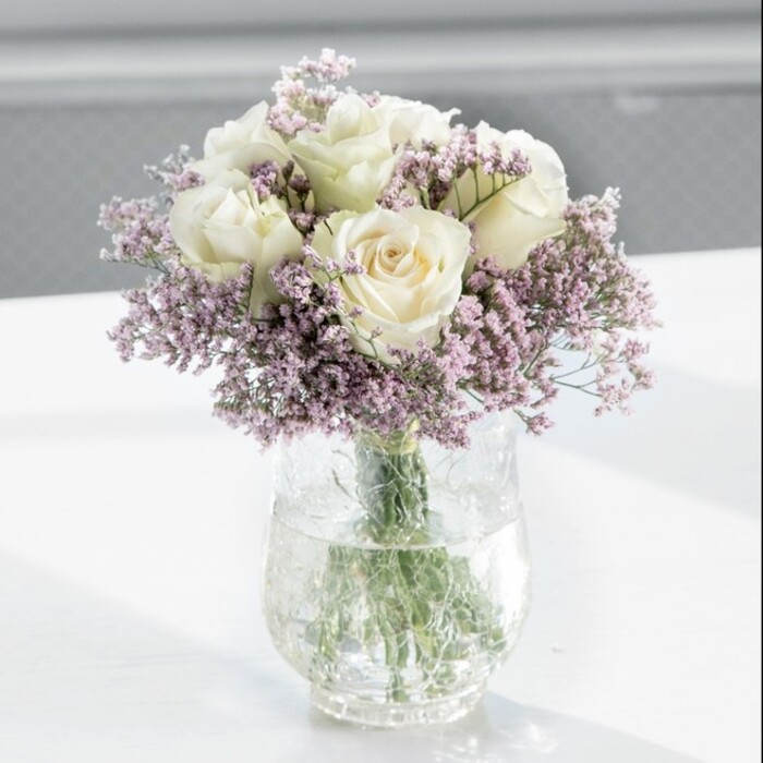 The image shows a classic, round bouquet dominated by lush white roses and framed by airy lavender statice, presented in a tall, cylindrical clear glass vase on a pristine white surface. Each rose is at that perfect stage of openness a florist looks for: the petals are layered and velvety, unfurling in soft spirals from a warm cream centre towards edges that fade into a gentle ivory. Their surfaces seem almost luminous under the soft natural light, recalling the pale tones of early morning skies over Mill Hill. Interlaced with the roses are generous clouds of tiny lavender-hued statice flowers, their branching stems and papery blossoms giving a light, cloud-like volume to the arrangement. These lilac accents form a delicate halo around the fuller rose heads, adding a fine, almost feathery texture that contrasts beautifully with the plush, rounded blooms. There is no heavy greenery to interrupt the harmony; instead, the eye moves from the creamy domes of roses to the misty purple sprigs and down through the glass to the orderly stems. The stems, neatly cut at an angle and gathered closely, stand in crystal-clear water, a sign that the bouquet has been freshly prepared, much like the arrangements sent out daily from independent florists around Mill Hill Broadway and along Flower Lane. Gentle daylight filters from an unseen window, the background softly blurred so that nothing distracts from the flowers themselves. The light casts subtle reflections on the tabletop and emphasizes the bouquet's serene palette. Overall, the mood is peaceful, graceful, and slightly nostalgic, making this arrangement suitable for a condolence bouquet delivered to a quiet home off Bittacy Hill, a refined centrepiece for a small Mill Hill anniversary dinner, or a thoughtful gift to bring soothing beauty to a contemporary office. The bouquet's restrained elegance and carefully balanced textures reveal the practiced hand of a florist who values both simplicity and emotional resonance.