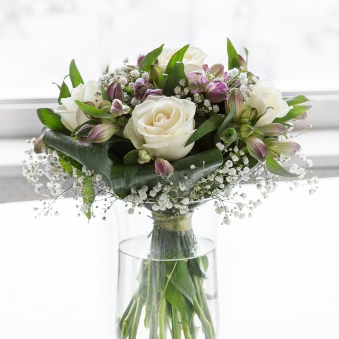 A refined floral bouquet stands gracefully in a clear, cylindrical glass vase, the water line revealing a tangle of neatly trimmed green stems that look as though they have just been cut fresh from an early morning market not far from Mill Hill Broadway. At the centre of the design, several white roses are in perfect, full bloom, their petals layered in soft, creamy spirals that catch the light and create a plush, almost cloud-like texture. These roses form the emotional heartbeat of the arrangement, radiating purity and quiet affection. Surrounding them are clusters of lavender and muted purple alstroemeria, their slender buds and partially open flowers introducing a gentle contrast. Some petals carry deeper violet veining, while others retain a cool, silvery-lilac tone that hints at dawn light over local gardens in Mill Hill East. Between the larger flowers, delicate sprays of white baby's breath weave through the composition, tiny blossoms perched on fine stems, adding a misty, ethereal quality that softens the overall shape. Lush greenery frames everything, from broad, dark green leaves that anchor the base to more delicate foliage that extends outward, giving the bouquet volume and movement without overwhelming its gentle palette. The natural daylight in the scene is soft and diffused, similar to light filtering through a sheer curtain or a north-facing window, making the background appear bright yet unobtrusive-a pale, almost luminous backdrop that allows every flower to stand in quiet clarity. The bouquet feels balanced and considered, crafted with the care you would expect from an experienced local florist who understands how to express sympathy, gratitude, or tender congratulations through form and colour. It is easy to imagine this arrangement resting on a dining table in a Mill Hill home, bringing a peaceful presence, a hint of delicate fragrance, and a sense of thoughtful connection to whoever receives it.