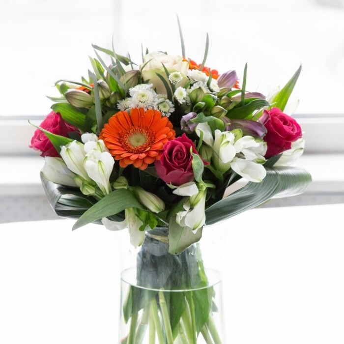 This stunning arrangement speaks to the artistry and heart of a true Mill Hill florist. Creamy peach gerberas surround the bouquet, forming a gentle ring of softness around luminous orange centers that glow like lanterns on a spring evening in Mill Hill Park. Those vibrant hues are balanced by lush magenta roses, their spirals of petals delivering a feeling of luxury and affection. Scattered throughout are ivory-hued alstroemeria and feather-light filler blooms that create layers and evoke the fresh tranquility of quiet roads like Daws Lane. The substantial aspidistra leaves, broad and confidently curled, embrace the bouquet's foundation, their robust green a calming anchor for the energetic florals above. The arrangement is bathed in natural light, each blossom's color magnified against a softly glowing background. Hints of rose and lily drift through the air, conjuring memories of local gardens after rain. This bouquet is perfectly suited for gatherings with friends, heartfelt milestones, or new ventures-every stem meticulously placed, capturing the optimism and camaraderie that define Mill Hill. The expert hands at Flowers Mill Hill ensure every piece is tailored for life's most meaningful moments.