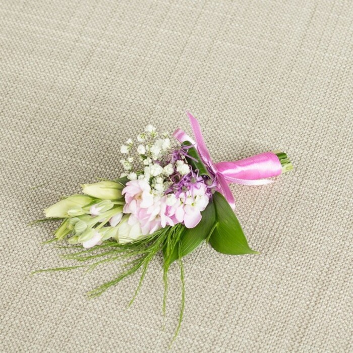 A small yet exquisitely detailed floral boutonniere or corsage is arranged on a neutral, finely woven linen cloth, the soft beige background allowing every floral nuance to stand out. At the heart of the design, several light pink, almost pastel blush blooms sit closely together; their many ruffled petals overlap like tiny, delicately folded silk fans, giving the central cluster a plush, romantic feel. Threaded throughout are numerous tiny white baby's breath flowers, each one a crisp, star-like dot that adds both texture and an airy brightness to the piece. Discreet flecks of lavender and deeper purple blossoms are tucked between the pink flowers and gypsophila, adding contrast and a refined hint of colour that keeps the palette interesting without overpowering its gentle mood. To the left, a short spray of creamy white to pale green buds, similar to young freesias or slender lilies, extends gracefully beyond the main cluster, their closed forms hinting at freshness and the unfolding of a special day. The entire arrangement is anchored by broad, emerald green leaves that form a smooth, glossy base. From beneath them, slender, thread-like green fronds arc outward, evoking the natural movement of grasses or fine asparagus fern, and lending the design a soft, garden-grown elegance. The stems at the base are firmly bound with a striking fuchsia satin ribbon, tied into a perfectly balanced bow, its sheen catching the light in gentle highlights. This carefully composed piece feels ideal for a groom or prom guest heading to a celebration in Mill Hill, perhaps after photos in Windsor Open Space, and could easily have been handcrafted by a local florist at Flowers Mill Hill for same-day delivery across the neighbourhood.