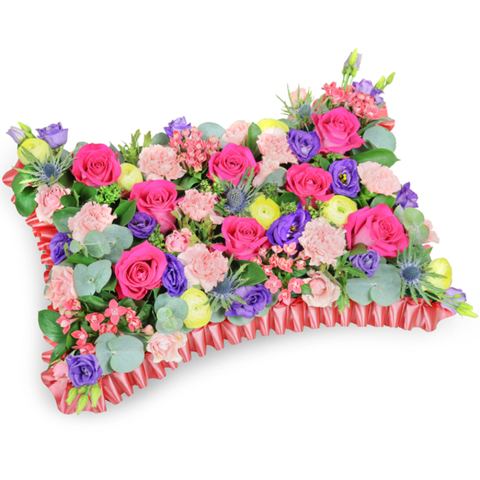 Captured from a slightly raised oblique viewpoint against a plain white surface, this pillow-shaped memorial arrangement presents a densely composed tableau of blooms, colour and texture that reads like a painterly still life. The surface is largely a chorus of hot fuchsia roses, their velvet petals arranged to create rhythmic pockets of depth, complemented by clusters of soft, pale-pink carnations whose frilly edges introduce a delicate relief across the top. Interspersed among these are royal purple lisianthus with graceful, bell-like forms, and cheerful lemon-yellow ranunculus that act as small beacons of light within the palette. Cool, spiky notes of silver-blue eryngium thistle give the piece structural interest, cutting across the blanket of softer florals with a wild, tactile energy. Underpinning the flowers is abundant greenery-rounded, silvery eucalyptus leaves alongside deeper green fillers-that provides contrast, scent and a restful backdrop. The arrangement sits upon an elegant ruffled red satin base; the pleated ribbon edge frames the composition and reinforces its cushion silhouette with a refined finish. Designed as a sympathetic tribute, the overall mood leans toward a celebration of memory: colours that speak of affection and stories well told, textures that invite calm reflection, and a scent profile where rose sweetness meets the cool of eucalyptus. This carefully balanced pillow would suit a funeral service, memorial, or graveside display in and around Mill Hill Village, offering both visual comfort and thoughtful craftsmanship reflective of local floral traditions.
