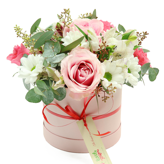 Set against a spotless white backdrop, a chic pastel pink hatbox filled with flowers appears as the single point of focus, much like a centrepiece waiting on a polished table in a quiet Mill Hill home. The box is round and cylindrical with a soft matte surface, wrapped by a narrow coral-red satin ribbon that is carefully tied into a petite bow at the front. From this bow, a cream-coloured gift tag marked with green, handwritten-style lettering hangs gently, hinting at a loving message from sender to recipient somewhere along Mill Hill Broadway or near Mill Hill Park. Emerging from the top of the box, the floral arrangement forms a generous, rounded dome of colour and texture. At its heart is an exquisite rose, its outer petals a gentle blush and its centre a deeper pink, unfolding in elegant spirals. Surrounding this focal bloom are several white daisy-like chrysanthemums, each with a vivid yellow-green centre that adds a bright, cheerful contrast and draws the eye across the bouquet. Delicate white alstroemeria interlace among these flowers, identifiable by their elongated petals and fine speckling, bringing an air of grace and lightness. Interspersed throughout are smaller deep pink and fuchsia blooms-likely spray roses or lisianthus-that offer rich, romantic notes against the calmer pastels. A generous mix of greenery completes the design: rounded, silver-green eucalyptus leaves give a contemporary, aromatic feel, while darker green foliage adds depth and structure. Subtle sprigs tipped with tiny buds or seed pods introduce a hint of wildness, as though the bouquet were partly gathered from a local garden. Bathed in even, soft light, every detail-from the satin sheen of the ribbon to the velvety rose petals-is clearly visible. The arrangement carries a sense of luxury and heartfelt intention, ideal for delivery across Mill Hill to mark birthdays, anniversaries, Mother's Day, or quiet moments of appreciation.