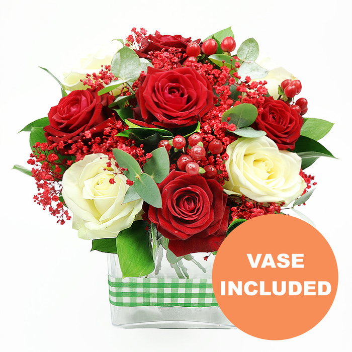 An opulent arrangement of red and cream roses is showcased against a spotless white background, the entire bouquet radiating a sense of thoughtful craftsmanship that would suit both modern apartments and traditional houses around Mill Hill and The Ridgeway. At the heart of the display, deep red roses open into lush, velvety rosettes, their rich tones suggesting romance, devotion, and heartfelt celebration. Between them, creamy white roses glow softly, their petals layered in gentle curves that temper the intensity of the red with a more tranquil, sophisticated note. Threaded throughout the roses are delicate sprays of tiny red filler flowers, akin to tinted gypsophila, which create a light, misty veil and soften the spaces between the larger blooms, giving the arrangement a slightly whimsical, festive feel. Scattered among the petals, glossy red hypericum berries cluster in small groups, their round forms and polished surfaces catching glints of light and adding both colour depth and tactile contrast. A generous collar of greenery frames the flowers, with dark green leaves lending structure and silvery eucalyptus sprigs bringing a cool, contemporary touch, reminiscent of the refined greenery found in well-kept Mill Hill gardens. The stems of the bouquet are gathered into a sleek, clear rectangular glass vase, its simple geometry enhancing the modern aesthetic while allowing the natural beauty of the green stems to remain visible. Wrapped around the centre of the vase is a green and white gingham ribbon, tied smoothly to impart a cosy, rustic charm that feels at home in family dining rooms near Mill Hill Broadway. In the lower right corner of the image, an eye-catching orange circular badge with white uppercase lettering declares VASE INCLUDED, clearly indicating that this romantic, ready-to-display arrangement arrives as a complete gift, ideal for anniversaries, Valentine's surprises, or simply bringing fresh elegance into a Mill Hill living space.