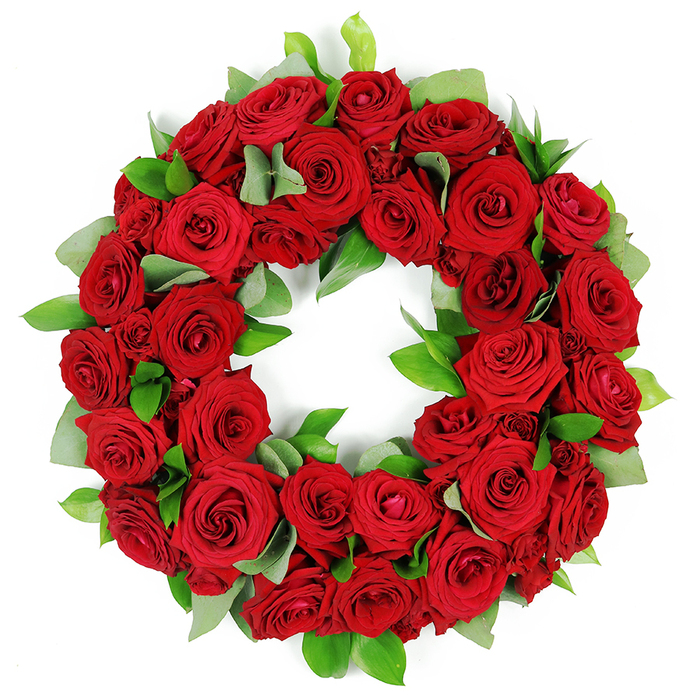 An artfully arranged red rose wreath, densely packed with dozens of velvety blooms, sits on a stark white background. The ring is perfectly formed, a continuous circle of crimson, with petals opening in stages from tight buds to full, layered rosettes. The colour is saturated, with subtle shading across the petals that creates depth and warmth. The greenery-eucalyptus-like broad leaves with a matte finish, and glossy, spear-shaped ruscus-weaves through the roses to provide texture and a bright green frame for the red. The piece reads as a solemn tribute, a classic symbol of love and remembrance appropriate for funerals, memorial services, or anniversaries. The arrangement is designed for impact: a full, three-dimensional presentation that invites close viewing, even against a white background that isolates its beauty. Crafted by a Mill Hill florist, it is a visible sign of sympathy that travels easily with delivery across Mill Hill and neighboring districts, ensuring the message of condolence reaches the right place at the right moment.