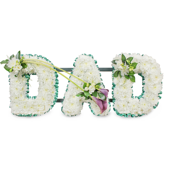 A beautifully composed funeral tribute spells out the word DAD in three large, three-dimensional block letters, the sort often chosen by families in Mill Hill to honour a much-loved father. Each letter is expertly covered in countless white chrysanthemums, or similar small button flowers, arranged tightly so that the petals touch and overlap, creating a thick, plush cushion of blooms. Their snowy whiteness conveys purity and remembrance, while the soft, rounded shapes of each flower head lend a gentle, comforting feel to the strong, upright capitals. Along the outer edge of every letter runs a rich emerald green ribbon, lightly ruffled, its satin surface catching the light and providing a striking outline to the flowers. The contrast between the cool white petals and the vivid green border evokes the calm of nearby green spaces in Mill Hill, such as the fields and tree-lined walks the area is known for. Behind the letters, faint hints of a dark frame reveal the sturdy structure that allows the tribute to stand upright as a single, unified piece.
The florist has added thoughtful touches of extra flowers and foliage to enhance the design without overshadowing the simple, powerful word. On the upper left arc of the first D, a dainty cluster of white freesia blossoms and rich greenery is nestled among the chrysanthemums, their slender buds and open bells adding depth and a light, sweet scent. From this point, two long, graceful green stems sweep diagonally across to the central A, visually linking the first two letters. At the lower right of the A, the stems culminate in an elegant, asymmetrical spray: two deep purple or maroon calla lilies, with their sculpted trumpet shapes, are arranged among more white freesia and fresh leaves, introducing a deeper, more contemplative colour accent. The final D carries a coordinating yet slightly more compact spray of white freesia and foliage on its upper right curve, echoing the first letter and bringing balance to the composition.
Set against a bright, uncluttered white background, this DAD tribute communicates love, respect, and profound loss in an understated but deeply moving way. It is a fitting choice for funerals, memorials, or cremation services throughout Mill Hill, offering families a visually clear and emotionally resonant way to say goodbye.