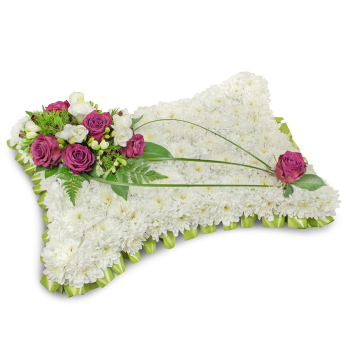 Set against a plain white field, this classic pillow-shaped funeral tribute is a study in peaceful simplicity: the entire cushion is tightly carpeted with dozens of pristine white chrysanthemum florets, their small, layered petals forming a uniform, cloudlike texture that reads as purity and rest. A neat, light-green satin ribbon is pleated around the lower edge, producing a delicate scalloped border that lifts and defines the silhouette. Dominating the top-left is a rich accent cluster of deep fuchsia roses - full, velvety blooms whose saturated color anchors the composition - interspersed with pale white freesias that lend a hint of fragrance and fragile contrast. Lush emerald foliage, including feathery fern fronds, frames the cluster and creates depth and shadow against the white blooms. From that cluster three slender, dark-green bear-grass stems arch elegantly across the face of the cushion, their long, sinuous lines guiding the eye toward the lower right, where a single matching fuchsia rose nestles among its own dark leaves to complete the visual conversation. The lighting is soft and even, casting a subtle shadow beneath the pillow and giving the arrangement a sense of calm weight. As a sympathetic tribute this pillow conveys repose and gentle remembrance - a thoughtful choice for funerals or memorials in Mill Hill Village, St. Paul's area, or local chapels near The Ridgeway - handcrafted with the care and quiet expertise expected from florists serving the Mill Hill community.