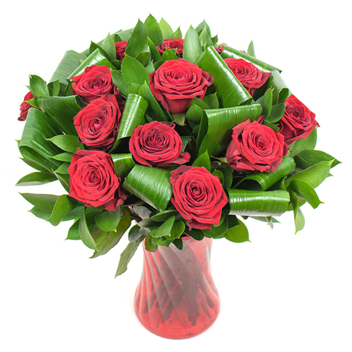 This close, composed view shows a generous cluster of deep crimson roses - twelve or more heads, each in full, perfect bloom with velvety, tightly wound petals that slowly unfurl toward soft outer edges. The roses read as almost burgundy in places, their colour amplified by gentle, even lighting that reveals subtle surface texture and the faint sheen of freshness. Interspersed among the blooms is an array of dark green foliage: compact, glossy leaves creating a dense, verdant bed and larger broad leaves that have been skillfully folded and rolled into elegant cylindrical shapes. Those rolled leaves act as living architecture, looping and spiralling to frame individual roses and to add modern structure to the classic form. The bouquet is anchored in a tall, translucent ruby-red glass vase with a slight ribbed twist that catches light in vertical streaks, complementing the deep tones of the roses and offering a graceful silhouette. Against a crisp white backdrop, the saturated reds and luminous greens pop with clarity, presenting the arrangement as both a statement of romance and a study in floral craftsmanship. You can almost sense the subtle perfume of red roses and the soft, tactile contrast between velvety petals and glossy leaves. For local context, imagine this arrangement gracing a candlelit table at a small celebration in Mill Hill Village or brightening an office in the Mill Hill Broadway area - a floral choice that reads as elegant, heartfelt, and carefully made by someone who knows the neighbourhood.