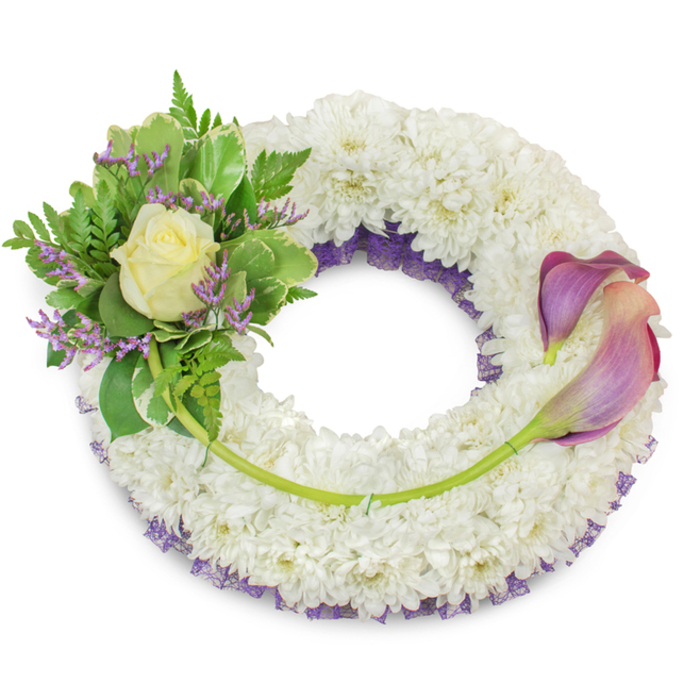 A large, dignified circular wreath built on a generous base of pure white chrysanthemums, the tightly clustered florets forming a textured, cloud-like surface that reads as tranquil and respectful. Beneath and between those white blooms, a ruffled layer of deep purple netting peeks out along the inner edges, giving the wreath dimensional contrast and a subtle, serif of colour. The upper-left segment is arranged with the precision of an experienced hand: a single cream or pale yellow rose opens gently at its centre, cushioned by rich green foliage that includes variegated leaves with pale margins and soft, lacy fern fronds. Tiny lavender-purple statice are dotted through this cluster, offering a restrained, powdery violet note that harmonises with the hidden purple under-layer.

On the right, two architectural calla lilies are laid to counterbalance the left cluster, their long, bright green stems curving across the lower-right arc and integrating into the chrysanthemum base for a seamless visual connection. Each calla shows a refined colour transition - darker purple at the edge fading into a blush or peach-pink nearer the throat - their smooth, sculptural forms creating movement and gravity. A single longer green stem travels across the bottom of the wreath, discreetly secured and leading toward the callas to bridge both sides. Photographed in soft, even light against a white backdrop, the arrangement suggests a considered, heartfelt tribute suitable for funeral services, graveside remembrances, or quiet memorials in the Mill Hill area near Mill Hill Park and Mill Hill Broadway.