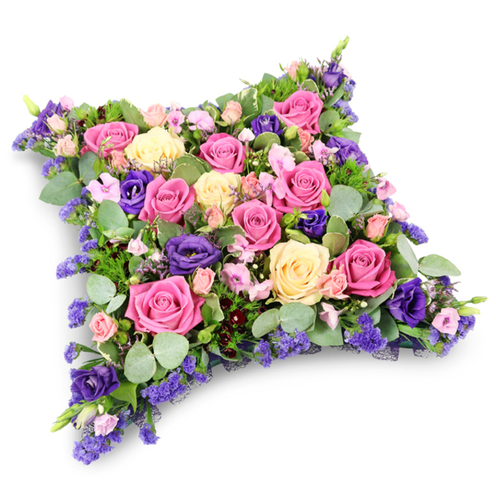 A thoughtfully composed floral tribute in the shape of a soft cushion dominates the image, its gently rounded rectangular form evoking the idea of a comforting pillow laid at a funeral or memorial in Mill Hill. The surface of the arrangement is densely packed with flowers, giving it a plush, almost padded appearance. Prominent among the blooms are large roses in shades of vibrant fuchsia, warm shell pink, and creamy pale yellow, many fully open and luxuriant, their spiralling petals forming layered rosettes that catch the light with a subtle sheen. Interspersed among these roses are elegant deep purple lisianthus flowers, whose velvety, ruffled petals introduce a sophisticated contrast and a dignified depth of colour. Filling the smaller spaces are delicate sprays of tiny lavender and lilac filler flowers, reminiscent of statice or limonium, creating a fine, airy texture and gentle clouds of soft pastel colour. Petite blush pink spray roses peek out between the larger blooms, adding a personal, affectionate touch. The flowers are underpinned and framed by an abundance of greenery: rounded, silvery-green eucalyptus leaves provide a cool, calming backdrop, while darker, more finely cut fern-like foliage adds structure and a whisper of woodland texture. The tribute is set against a stark white background, with a faint shadow beneath that subtly emphasises its three-dimensional depth and pillow-like presence. The overall effect is serene and tender, with the interplay of pinks, creams, and purples suggesting love, gratitude, and quiet reflection. It feels very much like a bespoke floral cushion created by local Mill Hill florists, suitable for delivery to a church, crematorium, or graveside as a graceful expression of condolence and enduring remembrance.