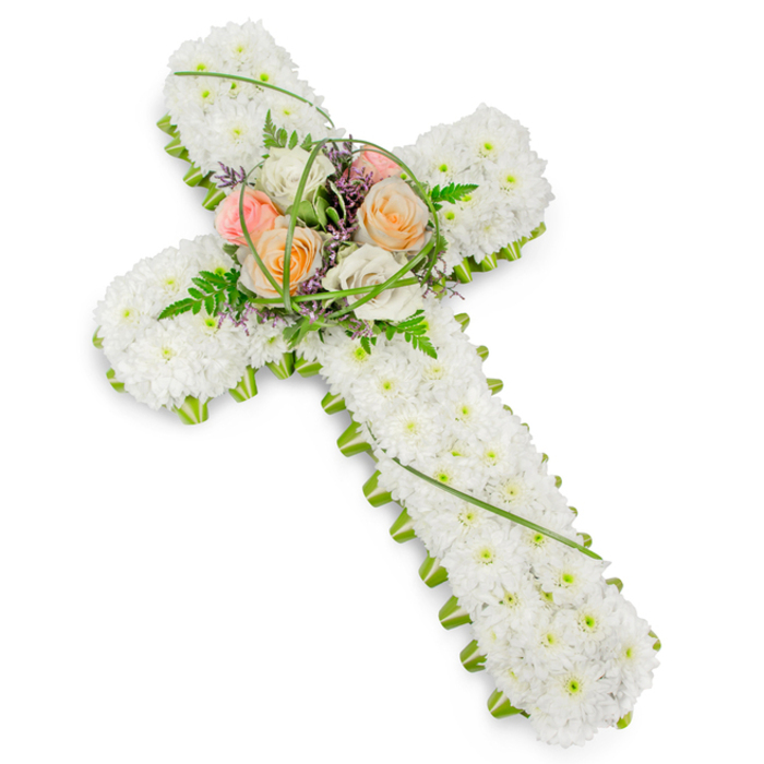 A reverent floral cross arrangement is shown in this image, its calm design laid out against a simple white background that keeps all attention on the flowers themselves. The cross is formed from densely clustered white chrysanthemums or similarly small blooms, creating a smooth, cushion-like surface that looks soft and inviting, as if it could gently cradle remembrance and prayer. The entire outline of the cross is accentuated by a narrow, bright green border of foliage, which runs along the arms and down the length of the base, clearly defining the shape much like the neat hedgerows and garden borders seen in homes around Mill Hill East. At the heart of the cross, a raised central spray of roses introduces a warmer, more personal note. The roses, fully open and lush, appear in a tender palette of soft peach, blush pink, and creamy ivory, with each bloom turned slightly in a different direction for a natural, garden-like feel. Around them, slender strands of bear grass loop and glide, while fern fronds and delicate greenery fill in the gaps, softening the outline of the focal point. Small, subtle flecks of lavender or mauve filler flowers glint between petals, adding a light wash of colour that enriches the design without overwhelming its peaceful mood. The light is gentle and even, ensuring that the whites stay soft and the greens feel fresh rather than stark. This floral cross feels appropriate for a funeral or memorial service in Mill Hill, a heartfelt tribute that embodies faith, comfort, and enduring affection from those left behind.