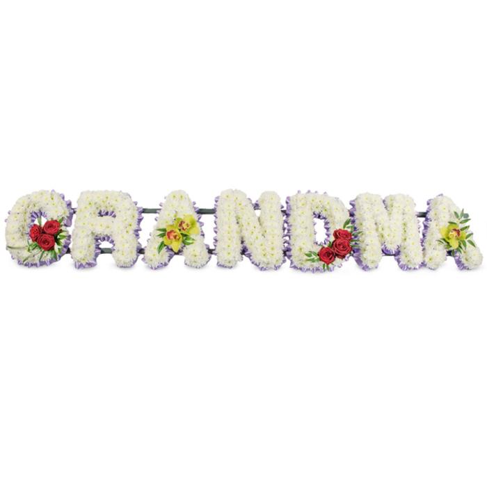 The photograph shows a carefully crafted GRANDMA flower tribute, created as a series of seven bold capital letters, each standing in a row against a spotless white background, much like it might appear on a stand at a quiet service in Mill Hill. The structure of each letter is formed from a metal frame, mostly hidden beneath a dense blanket of small white flowers. These blooms, likely chrysanthemums, are placed very close together, their rounded petals overlapping to form a thick, soft-looking surface that suggests both comfort and care. At the base of the letters, a trim of light lavender or pale purple flowers creates a neat outline, adding definition and a cool wash of colour that helps the letters stand out without overwhelming the simplicity of the white. This lilac edge feels calm and understated, echoing the gentle pace of the residential streets around Lawrence Street or the green open spaces nearby. Colourful accents are thoughtfully distributed across the word. On the G, near the lower left curve, three rich red roses form a tight cluster, framed by dark, glossy green foliage. Their deep red petals introduce a note of passion and devotion. The first A is highlighted with two yellow-green cymbidium orchids placed across the crossbar, their lightly speckled throats and unusual tone suggesting uniqueness and grace. The letters R, N, and D offer a quieter stretch of pure white petals, bordered only by the lavender trim, giving a sense of stillness at the centre of the tribute. On the M, another grouping of three crimson roses with leafy greens nestles at the lower right, mirroring the arrangement on the G and creating a pleasing visual balance. The final A repeats the pair of chartreuse orchids, completing the rhythm of colour that runs through the piece. All of the letters are aligned on a dark metal bar, discrete yet sturdy enough to support the tribute during transport and display at a chapel, cemetery, or crematorium in Mill Hill. The entire arrangement conveys a blend of purity, warmth, and personal remembrance, offering a gentle floral message of love for a much-missed grandmother.