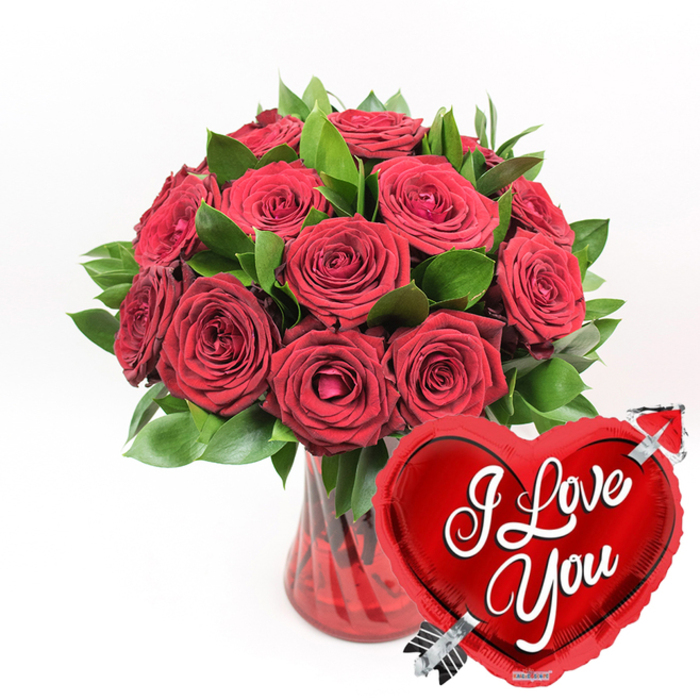 This photograph presents a timeless romantic scene: a generous bouquet of deep crimson roses, freshly arranged in a ruby-edged glass vase, paired with a radiant heart balloon that declares love in unmistakable terms. At the centre, around a dozen or more long-stemmed roses have been clustered together into a lush, rounded bouquet. Some of the blooms are fully open, their petals unfolding in intricate spirals that give a sense of volume and softness, while others remain semi-closed, their outer petals gently curled, suggesting youth and continuity. The roses are surrounded and supported by vibrant green foliage, the leaves smooth, slightly waxy, and shaped to fan out, creating a layered backdrop that makes each flower head stand out. The colours feel intensified, as though seen in natural daylight streaming into a Mill Hill home near Daws Lane. The arrangement is cradled in a clear ruby-red glass vase with subtle vertical fluting; the ribbed texture catches the light in thin lines, giving the vase a refined, tactile look. Through the translucent red glass, glimpses of the sturdy green stems and crystal-clear water are visible, grounding the bouquet and hinting at its freshness. In the lower right foreground, overlapping the vase and edging into the roses, a metallic red heart-shaped foil balloon adds a playful yet deeply sentimental touch. The balloon's shiny surface reflects light in broad highlights, making the shape appear full and buoyant. Across its centre, bold white cursive writing reads I Love You, while a silver arrow motif cuts diagonally through the heart, evoking Cupid and love struck at first sight. The entire composition sits against a clean, white backdrop, free from distractions, allowing the rich reds, vivid greens, and reflective silver to command attention. It's an image that feels made for a romantic delivery in Mill Hill Broadway, a proposal dinner at home, or a heartfelt anniversary message shared across North West London.