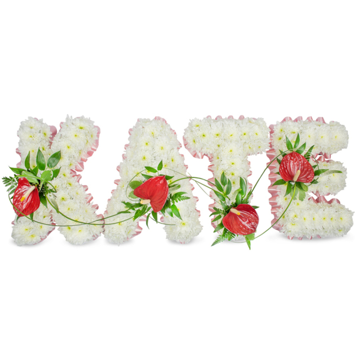 This bespoke funeral tribute spells 'KATE' in prominent white capital letters, each letter carefully built from a dense, uniform carpet of small white spray chrysanthemums that create a soft, cloud-like surface. The chrysanthemums are packed closely to produce an even, tactile texture, appearing almost cushiony to the touch and diffusing a delicate, green-floral scent that feels understated and comforting in a quiet service. A pale pink, ruffled ribbon runs along the outer contours of every letter, its pleated edge providing a refined, subtle frame that warms the overall palette without overwhelming it. Set into the white base are vivid clusters of red anthurium blooms: glossy, heart-shaped spathes with their characteristic spadices that offer a striking, heartfelt contrast to the white. Around these focal blooms sits a selection of deep green foliage - broad glossy leaves, feathery fern tips, and fine trailing tendrils - carefully interlaced to give depth and a gentle sense of movement between letters. The tendrils and vines sweep across the composition, visually binding the separate letters into a single, cohesive tribute. Presented against a bright white background with a soft drop shadow beneath each character, the arrangement reads as both elegant and substantial. A Mill Hill florist would note how this piece suits chapel viewings, wakes near Mill Hill Village, or remembrance gatherings: its clean lines, considered textures, and modest ribbon detail convey respect, warmth, and enduring memory with quiet craftsmanship.