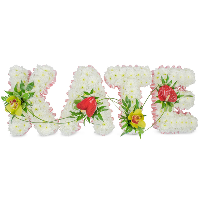 A carefully composed floral name tribute spells out KATE in four substantial capital letters, arranged side by side on a simple white background that enhances their clarity and presence. Each letter is filled edge-to-edge with pristine white chrysanthemums, their densely packed, multi-petalled heads creating a padded, textured surface that appears soft to the touch yet precisely shaped. Tracing the contours of the K, A, T, and E is a narrow band of pale pink ribbon, gently gathered to form a ruffled border that introduces a light, romantic note and emphasises the clean outline of each letter. Within this gentle framework of white and pink, the florist has set key accent flowers that provide colour, structure, and emotional resonance. On the left bar of the K and clasped near the foot of the T, yellow-green cymbidium orchids with warm, speckled reddish-brown throats emerge from clusters of lush green foliage, adding brightness and a sense of refined luxury. The A and the E each feature a striking red anthurium bloom, heart-shaped and glossy, whose waxy texture and deep colour stand out vividly against the soft chrysanthemum base, echoing themes of love, remembrance, and courage. Fine fern fronds and sleek green leaves surround these focal blooms, while slender tendrils arc and curl from one letter to the next, gently linking the name as though a single strand of greenery ties KATE together. The overall impression is of a thoughtful, bespoke funeral arrangement, the sort of personalised tribute a family in Mill Hill might choose for a funeral, memorial, or celebration of life when they wish to see their loved one's name expressed in flowers with both clarity and heartfelt grace.