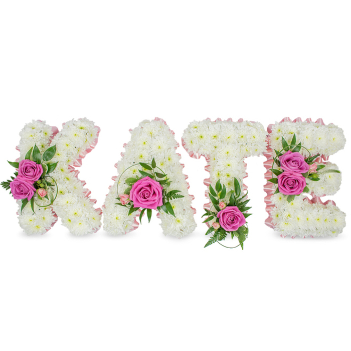 A carefully composed, personalised floral name tribute spells out KATE in large, freestanding letters, each formed from a dense carpet of small, immaculate white chrysanthemums that create a plush, cloud-like texture and flawless, continuous surface. Soft pink satin ribbon traces the inner shapes of each letter, providing an understated contrast that refines the edges and adds a whisper of colour against the white base. Attention is drawn to vivid hot pink roses placed with deliberate rhythm: two roses on the left arm of the K among smaller spray roses and spiral grass, a single prominent rose held in the lower centre of the A with surrounding light-pink buds, one bright rose anchoring the base of the T accompanied by fern-like fronds, and a pair of hot pink roses on the right of the E accented by variegated green leaves. Interspersed greenery-glossy ruscus, feathery fern tips and slender eucalyptus-creates depth, cools the palette and imparts a subtle, fresh fragrance beneath the sweeter rose notes. The set is staged against a clean white background so shadows define the three-dimensional form, making the letters stand clear in a chapel, crematorium or funeral reception. As a thoughtful tribute offered by a Mill Hill florist, this arrangement reads as intimate, reverent and handcrafted-an elegant expression of love and remembrance suitable for memorial services, life celebrations or as a personalised funeral tribute for someone dearly missed in the local community.