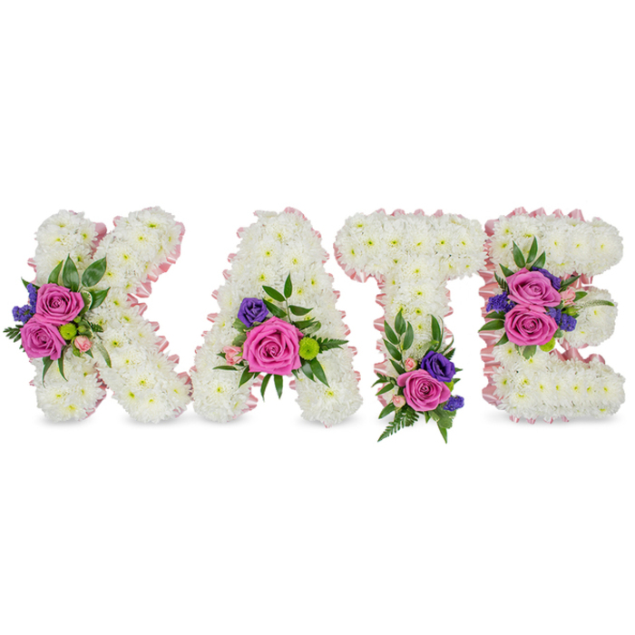 A classic memorial arrangement spelling the name KATE in four freestanding letters, each built from a thick, snow-white carpet of chrysanthemums that give a full, cushiony texture and a sense of calm. The letters are edged with a pale pink satin ribbon that traces the contours and inner voids, the ribbon's faint sheen lending a gentle, almost candlelit glow to the composition. Strategic clusters of colour punctuate the white: bold fuchsia-pink roses anchor the groupings, accompanied by softer pastel pink spray roses and slender stems of deep purple or blue accents-likely statice or small lisianthus-that add quiet contrast and a refined silhouette. Eucalyptus leaves and fine fern fronds weave through the clusters, their muted greens and airy shapes softening the overall profile and introducing a subtle herbal scent beneath the sweeter rose notes. Placement is thoughtful and balanced-the K holds a concentrated cluster on its diagonal stroke, the A carries blossoms near its lower right curve, the T receives a grounding spray close to its base, and the E is decorated with two distinct clusters on the center bar and bottom arm-each position chosen to honour the name with dignity. Intended as a bespoke sympathy or funeral tribute, this piece speaks with gentle authority and tenderness. Prepared locally in Mill Hill by Flowers Mill Hill, it suits services across the village and nearby chapels around The Broadway, offering a personalised and consoling expression of remembrance.