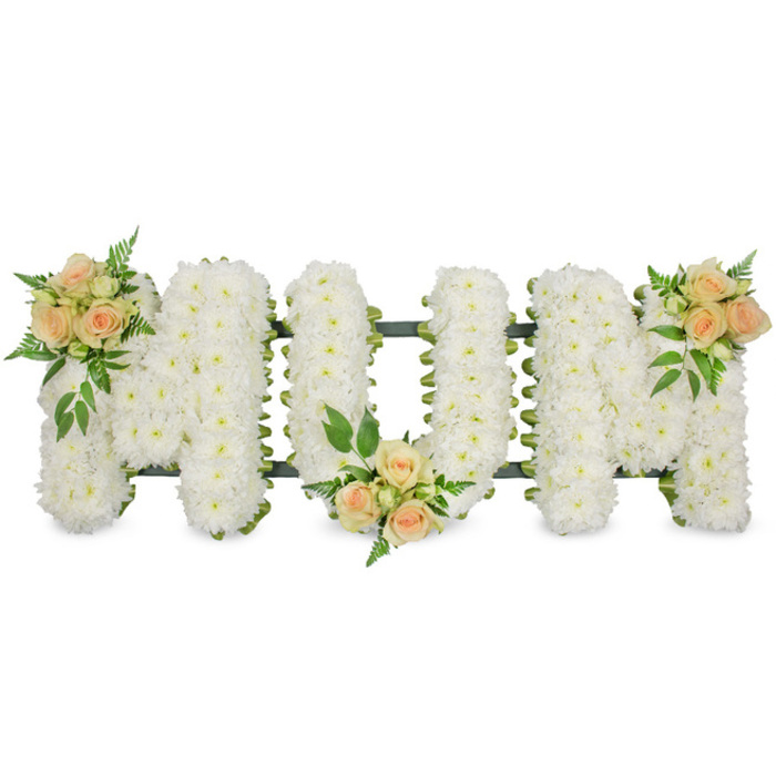 This photograph shows a classic MUM letter tribute for a funeral, laid out in a clear horizontal line and captured against a spotless white background that gives it a peaceful, uncluttered setting. Each of the three uppercase letters is completely filled with small white spray chrysanthemums, packed so closely that they form a continuous, cloud-like layer of petals. The chrysanthemums' tiny, rounded blooms create a soft and almost cushiony texture, suggesting comfort, purity, and serenity. Peeking through around the sides is a slim dark green ribbon and the supporting frame, outlining the letters with a neat border that enhances their shape and provides a touch of contrast. Resting delicately upon this white floral base are three sprays of roses in gentle peach and blush pink shades. One sits on the upper left arm of the first M, another is cradled along the lower curve of the U, and the final cluster crowns the upper right arm of the second M, resulting in a subtle visual rhythm that draws the eye across the word. These rose accents include both open blossoms and smaller buds, framed with feathery ferns and lush green leaves that add layers of texture and a natural, garden feel. The arrangement carries a mood of quiet reverence and affectionate remembrance, ideal for honouring a mother at a funeral service or cremation. It is easy to imagine this tribute being delivered to a Mill Hill chapel or resting place, bringing a gentle presence of beauty and solace as family and friends gather to celebrate a life that meant so much.