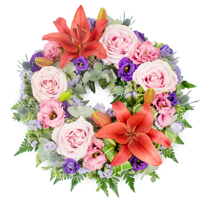This image presents a meticulously crafted circular funeral wreath, laid flat against a pristine white background so every detail of the floristry is clearly visible. The wreath's form is a perfect ring, symbolising continuity, and is anchored by two prominent coral-orange Asiatic lilies, one placed near the upper curve and the other toward the lower side. Their petals are fully open, gently curling back to reveal pronounced stamens that add depth and a touch of drama. Surrounding these focal lilies is a profusion of lavender-pink roses, each one at its peak of bloom, with layers of velvety petals spiralling inward to form a soft, romantic centre. Interspersed throughout are eustoma, or lisianthus, in both rich purple and pale pink-lavender shades; their ruffled, cup-shaped blossoms introduce a flowing texture that contrasts with the more structured roses. Between the larger blooms, small spiky heads of blue-grey eryngium pop up, providing a cool, architectural accent, while delicate sprays of lavender-blue statice lace through the design, adding lightness and a misty veil of colour. The base of the wreath consists of abundant mixed foliage: smooth, rounded eucalyptus-like leaves, fine fern fronds, and other varied greens, all contributing to a dense, verdant foundation. Near one of the lilies, bright green folded leaves, likely aspidistra or Ti, are tucked in with precision, showing the careful craftsmanship associated with Flowers Mill Hill. This thoughtful wreath would suit a chapel service near Mill Hill Broadway, a quiet ceremony by Arrandene Open Space, or a memorial gathering at a local cemetery, conveying a message of peaceful remembrance, respect, and enduring love.
