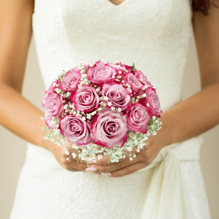 Against the soft light filtering through a Mill Hill workshop window, this intimate image presents a bride, captured from mid-chest down, tenderly embracing a rounded bouquet awash in gradients of pink. The arrangement features a dozen perfectly formed roses, each bloom transitioning seamlessly from delicate blush in the center to a luxuriant magenta at the outer rim, echoing the riot of colours seen in Mill Hill's springtime gardens. Wisps of baby's breath intermingle between the roses, spilling over like delicate clouds and offering a gentle, dreamy foil to the vibrant flowers. Hints of fresh foliage peek out at the top, melding harmoniously with the bridal white of the gown. The bride's hands-warm with a natural tan and nails polished in soft neutrals-gently cup the bouquet, the fabric of her sleeveless, lace-adorned dress gathering with a sash just above her wrist. The floral lace motif and deep V-neckline imbue the whole look with classic romance, the kind celebrated at local Mill Hill venues and along bustling Broadway. The background is softly blurred and neutral, allowing each floral detail to stand out with clarity and grace. This scene encapsulates the care and creativity of local florists, evoking the joy and excitement of a wedding day in NW7-an image destined to inspire those dreaming of walking down the aisle surrounded by timeless beauty and heartfelt blooms.