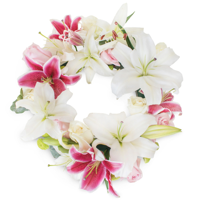 Inspired by the peaceful, green expanses of Mill Hill, this elaborate sympathy wreath artfully combines grand blooms with gentle elegance. A seamless ring of pristine white Oriental lilies forms the base, their broad petals curling outward like soft ribbons, each open blossom revealing pale green pistils and subtle golden stamens that signal innocence and hope. Contrasted beautifully are vivid fuchsia-pink lilies, their speckled petals glimmering with saturated magenta against the surrounding white, bringing striking energy to the arrangement. Between these commanding lilies rest bunches of creamy white and blush pink roses, some in an unfurling state, others still tightly budded, imbuing the circle with layers of texture and quiet romance. Sprigs of muted green foliage-perhaps inspired by the greenery in Mill Hill's own parks-peek gently over and between the blooms, adding dimension and a suggestion of local freshness. Isolated on a crisp white background, the wreath's continuous form suggests completeness, with each detail rendered in vivid clarity. The careful touch of Flowers Mill Hill is evident, from the harmonious blending of colours to the precision of placement, ensuring a composition worthy of memorial occasions at local chapels or as a cherished condolence gesture throughout Mill Hill. This floral wreath stands as both an artistic centrepiece and a comforting token, representing the circle of life and the nurturing spirit of NW7's community.