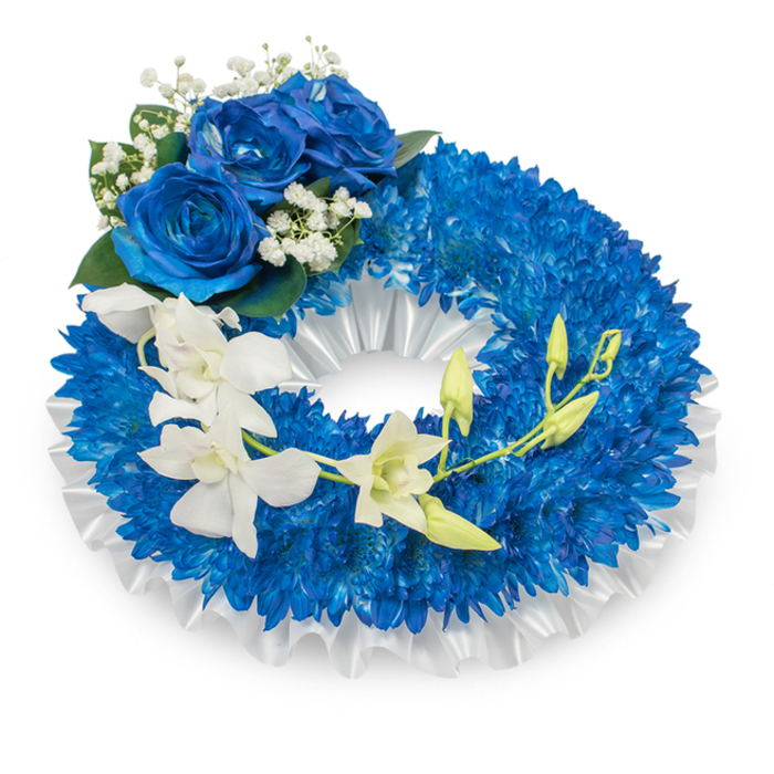 Presented against a clean backdrop, this circular sympathy wreath combines saturated blue and pure white in a composition that feels both formal and gently comforting. The main body is densely covered with countless small, intense royal-blue chrysanthemum blossoms whose tightly packed, almost pompon-like heads create a plush, textural ring. A pleated white satin ribbon lines the inside and subtly shows at the outer edge, its soft folds giving the wreath a refined frame and a slight, elegant lift. At the upper-left curve sits a cluster of three full blue roses, their thick, velvety petals opening in a deep sapphire that harmonises with the chrysanthemum base; these roses are accented by tiny sprays of white baby's breath and deep green leaves that provide contrast and breathe around the focal blooms. From the mid-left segment a sweep of clean white orchids - with the familiar flat faces and delicate lips of Phalaenopsis or the slender form of Dendrobium - drifts downward toward the lower right, the blooms appearing almost luminous against the dark blue. Intermingled along that flowing stem are lime-tinged green buds at various stages of opening, signalling freshness and ongoing life within a memorial context. Bathed in soft, cool light, the arrangement reads as a composed tribute suitable for services in Mill Hill Village or small local chapels, conveying peace, reverence, and a careful, considered hand in floral craftsmanship.