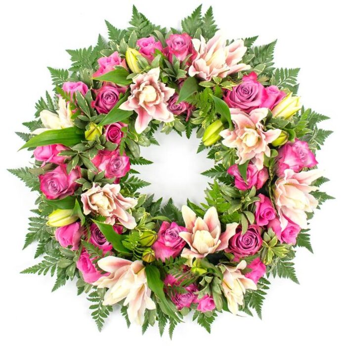 This image shows a meticulously crafted circular sympathy wreath, centred against a luminous white backdrop that allows its colours to stand out as clearly as if seen in soft chapel lighting. The wreath's structure is lush and full, composed so that flowers and foliage blend into an unbroken circle of remembrance. Dominating the design are roses in a refined pink palette: intense magenta blooms, vibrant fuchsia heads, and softer blush roses appear in different stages of opening, from small, tight buds to expansive flowers whose petals curl outward in layers. Intertwined among them are luxurious double lilies in creamy white and delicate pastel pink, some with a suggestion of peach or deeper pink near the petal tips. Their broad petals give the wreath a star-like rhythm, and several lime-green lily buds lend height and freshness, as if the arrangement has only just been completed. Around and beneath this floral band sits an abundance of greenery. Fern fronds, with their characteristic feathery texture, outline the outer edge like a soft green lace, while broader, shiny leaves-some slightly variegated-fill the inner spaces, making the wreath appear deep and substantial rather than flat. The white background eliminates any visual noise, so each petal, leaflet, and bud can be distinctly imagined. The atmosphere is serene and respectful, perfectly suited to a funeral or celebration of life. It is the kind of wreath families in Mill Hill might choose for a farewell at a local church, or to be delivered to a service near Mill Hill Broadway, trusting that the floral tribute will reflect their affection and respect with quiet, polished beauty.