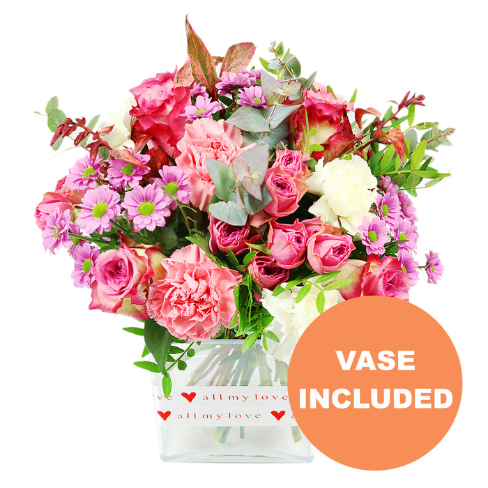A vibrant, thoughtfully composed floral arrangement fills this image, featuring a rounded bouquet standing tall in a clear, square glass vase with softly rounded corners, set against a bright white background. The dominant impression is one of romantic pinks and gentle pastels, arranged with the practiced touch of a local Mill Hill florist. At the centre, multiple roses in rich fuchsia and tender blush-pink tones showcase velvety petals layered in spirals, some blooms fully open and others still unfolding. Interlaced with the roses are large, ruffled carnations in hues of coral, peach, and creamy white, their soft frills adding dimension, movement, and a sense of abundance. Scattered throughout the design are petite daisy-like chrysanthemums in lavender and light purple, punctuated by striking lime-green centres that provide an uplifting brightness, much like a sunlit view from Mill Hill Broadway on a clear day. The bouquet is framed by an assortment of greenery: rounded, silvery-green eucalyptus leaves, darker glossy foliage, and the occasional touch of reddish-brown leaves, which bring a gentle, almost autumnal warmth and emphasise the depth of the arrangement. The glass vase itself is encircled near the base by a white decorative band, printed with small red hearts and the phrase all my love in fluid, romantic script, making this design especially apt for anniversaries, Valentine's surprises, or heartfelt thank-you gifts delivered across Mill Hill and the surrounding streets. In the lower right corner of the image, a bright orange circular graphic announces VASE INCLUDED in bold white capital letters, clearly signalling that this is a complete, ready-to-display bouquet suited to any Mill Hill home, from apartments near the station to houses along The Ridgeway.