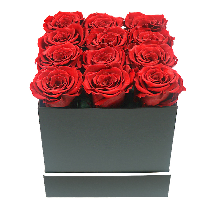 In this photograph, a chic, minimalist rose box arrangement is presented with the polished calm of a Mill Hill boutique. A square box sits centrally against a spotless white background, its matte black surface giving off a soft, muted look rather than shine. The lines of the box are crisp and geometric, exuding a contemporary style often favoured in the elegant homes around Mill Hill and The Ridgeway. Around the lower part of the box, a thin, bright white band wraps all the way around, providing a gentle but unmistakable contrast. Emerging from the open top are twelve lush red roses, their heads gathered so closely that they form a nearly seamless carpet of petals. Arranged in a neat 3x4 grid, the blooms appear almost identical in size and stage of opening, each one fully unfurled with layered, velvety petals. The colour is a rich ruby red, deep and romantic, with the inner spirals of the roses slightly darker, adding depth and dimension to the overall effect. The flowers rise slightly above the rim of the box, giving the impression of fullness and abundance without any greenery or filler to distract from their beauty. The pure white backdrop isolates the arrangement and makes the interplay of red and black especially striking. Soft, even lighting falls across the composition, avoiding harsh contrasts while gently emphasizing the curled edges of the petals and the sharp corners of the box. The overall atmosphere is one of refined romance and considered luxury, perfectly suited for a lavish Valentine's gift or a special anniversary surprise delivered to a quiet street near Mill Hill Broadway or overlooking Arrandene Open Space.