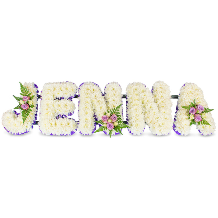 A tender, name-shaped floral tribute spells JENNA in large, three-dimensional capital letters crafted from a dense swathe of pristine white chrysanthemums or similar delicate florets, arranged against a spotless white background to emphasize purity and calm. Each letter has a soft, cloud-like volume created by tightly packed petals, while a vivid purple trim-either a narrow ribbon or a row of smaller blooms-traces the inner edges to give each character definition and a quietly regal accent. Three small floral groupings punctuate the composition: soft pink and lavender spray roses, alongside a few pale cream or buttery yellow roses, sit among fresh, dark-green fern fronds at the upper curve of the 'J', the lower centre of the first 'N', and the upper right of the 'A'. Slim, dark structural bars subtly join the letters, ensuring the piece reads as a single, composed tribute suitable for placement in a church, funeral home, or beside a cremation table. The fragrance is gentle and consoling-cool chrysanthemum notes softened by the sweet trace of roses-while studio lighting brings out the contrast between the immaculate white and the purple edging. As a Mill Hill florist familiar with local services near Mill Hill Broadway and family gatherings at nearby green spaces, I picture this arrangement positioned where mourners can read the name easily from the pews, and approach to see the delicate craftsmanship up close. The overall mood is one of respectful remembrance: elegant, handcrafted and quietly consoling.